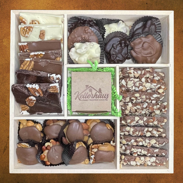 Wooden tray with 5 compartments containing buttercrunch, bark, chocolate caramel nut clusters and chocolate nut clusters, and a box with chocolate covered caramels in the center.