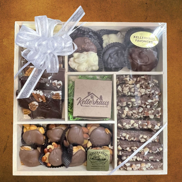 Wooden tray with 5 compartments containing buttercrunch, bark, chocolate caramel nut clusters and chocolate nut clusters, and a box with chocolate covered caramels in the center.