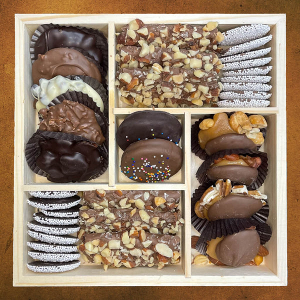 Wooden tray with 5 compartments containing butter crunch, nonpareils, chocolate caramel nut clusters and chocolate nut clusters, and a box with chocolate covered Oreos in the center.