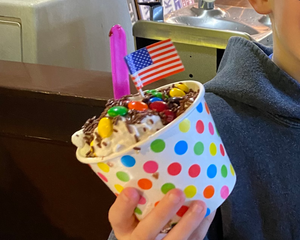 Child holding a colorful ice cream sundae with a small American flag, standing in a casual setting.