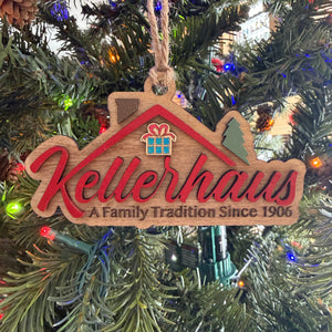 A wooden house-shaped Christmas ornament hanging on a decorated Christmas tree. The ornament is light natural wood with red script lettering that reads 'Kellerhaus' and smaller text below reading 'A Family Tradition Since 1906'.