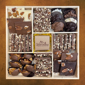 Wooden tray with 5 compartments containing butter crunch, bark, chocolate caramel nut clusters and chocolate nut clusters and a box with chocolate covered caramels at the center